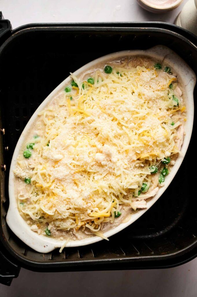 Oval baking dish with uncooked Air Fryer Chicken Tetrazzini, topped with shredded cheese and peas, placed inside an air fryer basket.