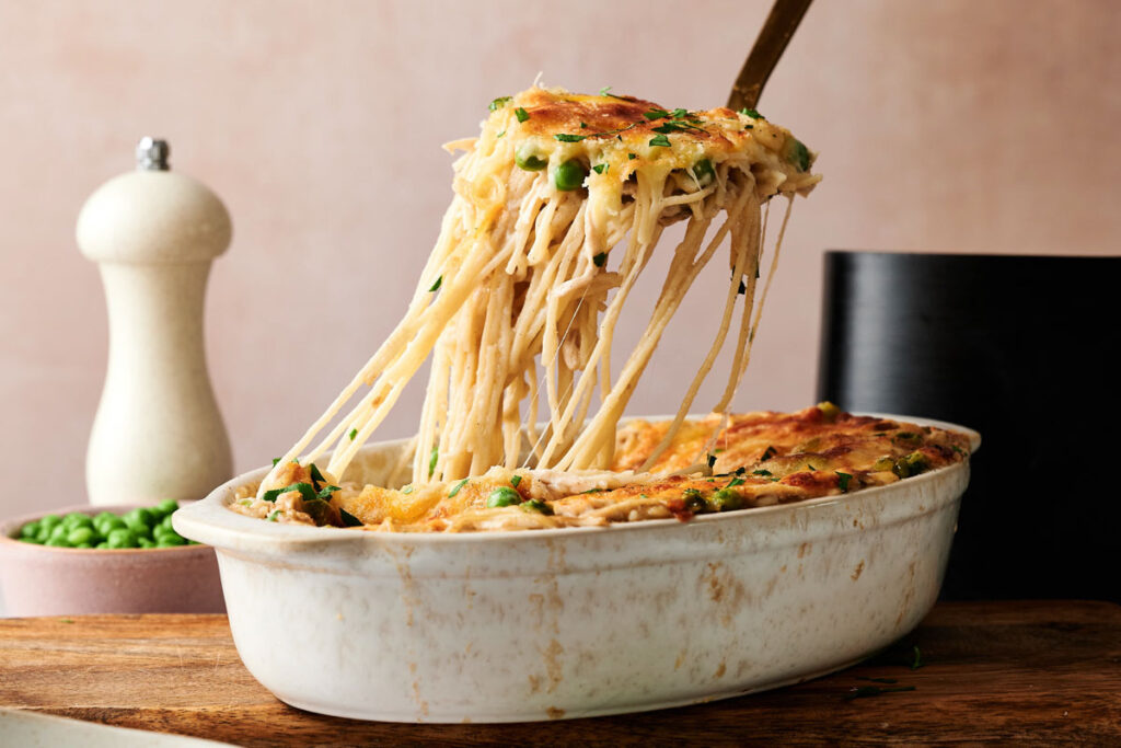 A serving of Air Fryer Chicken Tetrazzini is being lifted from a white casserole dish, showing melted cheese and herbs on top, with a pepper grinder and a bowl of peas in the background.