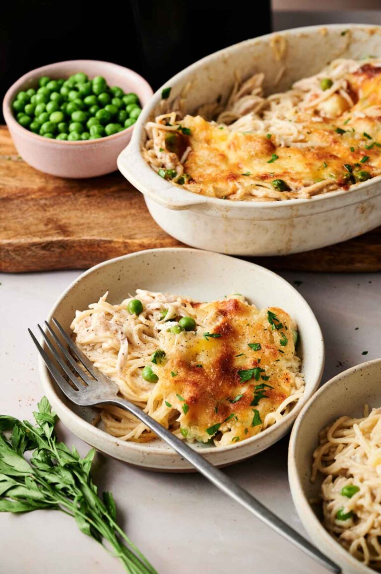 A bowl and baking dish of baked spaghetti casserole with peas and melted cheese, reminiscent of Air Fryer Chicken Tetrazzini, served with a fork; a small bowl of green peas and parsley garnish nearby.