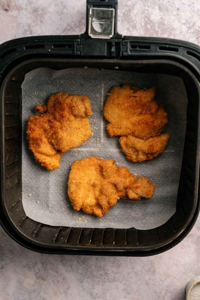 Three breaded chicken cutlets, reminiscent of Air Fryer Crispy Chicken Thighs, rest on parchment paper inside an air fryer basket.