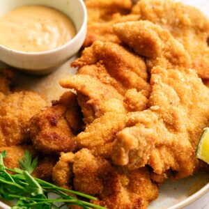 A plate of breaded and fried chicken tenders, inspired by Air Fryer Crispy Chicken Thighs, served with a small bowl of dipping sauce and a garnish of fresh parsley and a lime wedge.