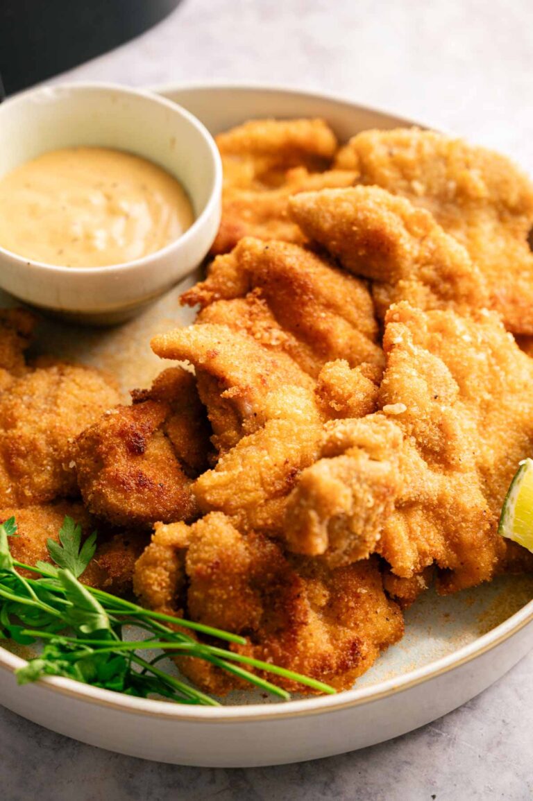 A plate of breaded and fried chicken tenders, inspired by Air Fryer Crispy Chicken Thighs, served with a small bowl of dipping sauce and a garnish of fresh parsley and a lime wedge.
