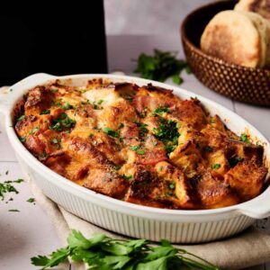 An Air Fryer Eggs Benedict Casserole, topped with chopped herbs, sits in a white dish on a beige cloth, with parsley and a basket of bread in the background.