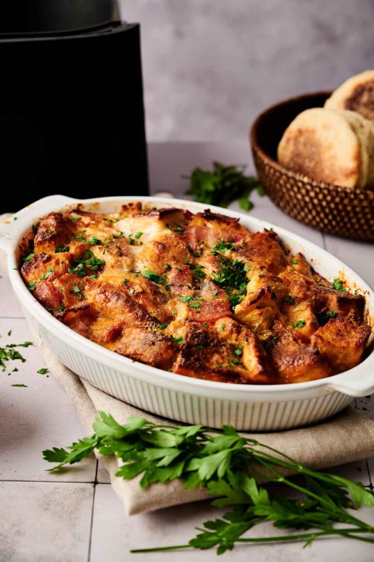 An Air Fryer Eggs Benedict Casserole, topped with chopped herbs, sits in a white dish on a beige cloth, with parsley and a basket of bread in the background.