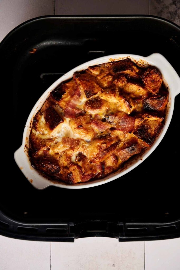 Oval white baking dish filled with golden-brown Air Fryer Eggs Benedict Casserole, showcasing toasted bread cubes and melted cheese, placed inside a black air fryer basket.