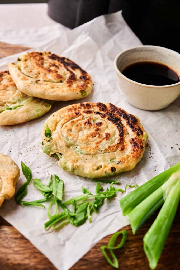 Three Air Fryer Scallion Pancakes on parchment paper, garnished with chopped green onions, served beside a small bowl of soy sauce.