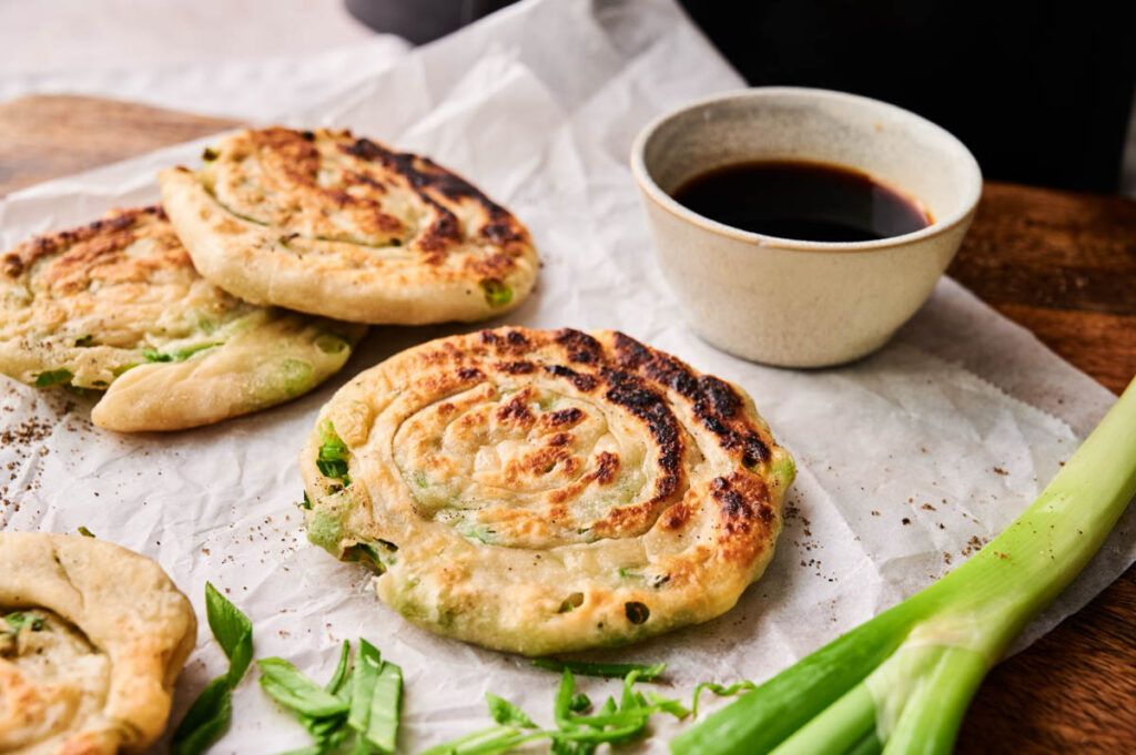Three Air Fryer Scallion Pancakes on parchment paper sit next to a bowl of dipping sauce and fresh green onions.