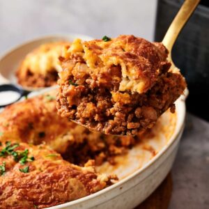 A serving spoon lifts a portion of golden, crispy Air Fryer Sloppy Joe Casserole, reminiscent of classic shepherd&rsquo;s pie, from a white casserole dish.