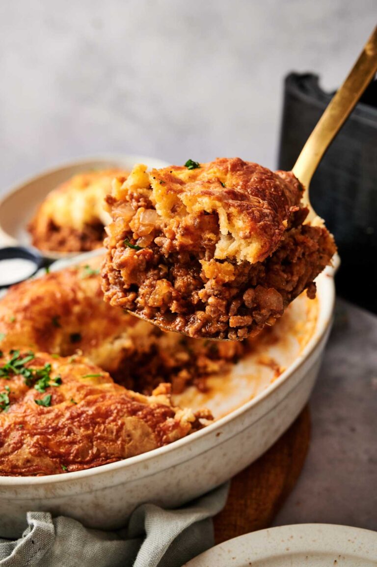 A serving spoon lifts a portion of golden, crispy Air Fryer Sloppy Joe Casserole, reminiscent of classic shepherd’s pie, from a white casserole dish.