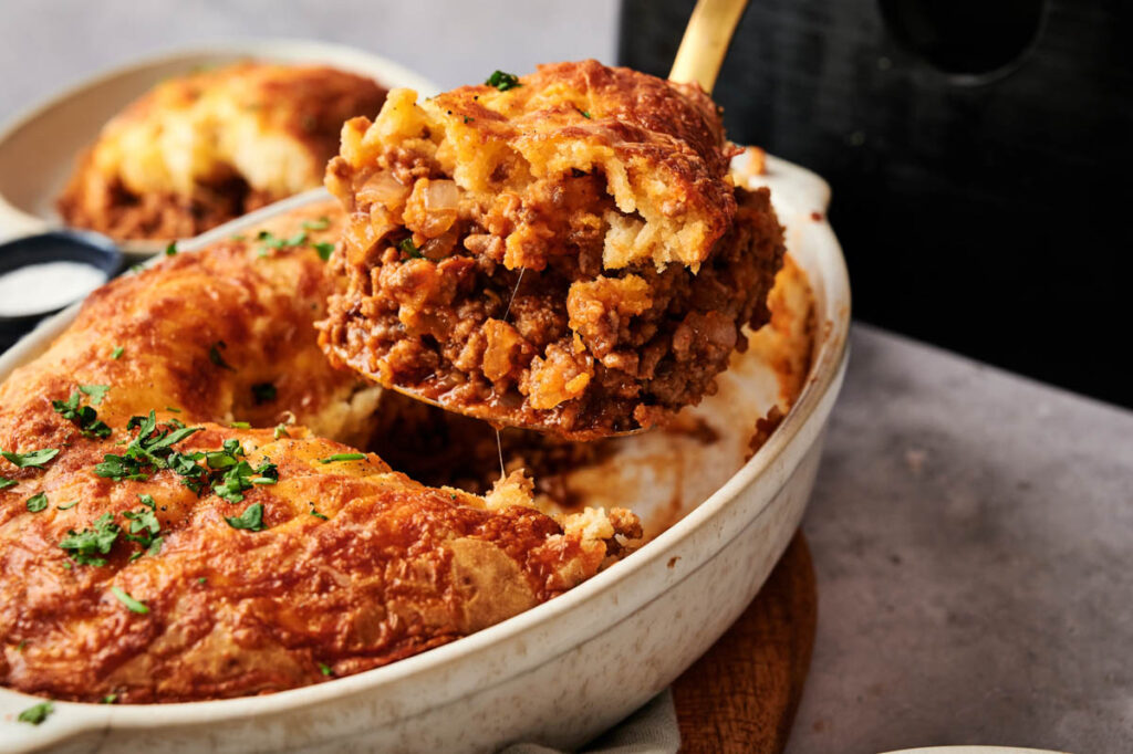 A serving spoon lifts a portion of Air Fryer Sloppy Joe Casserole, topped with melted cheese and garnished with chopped parsley, from a white baking dish.
