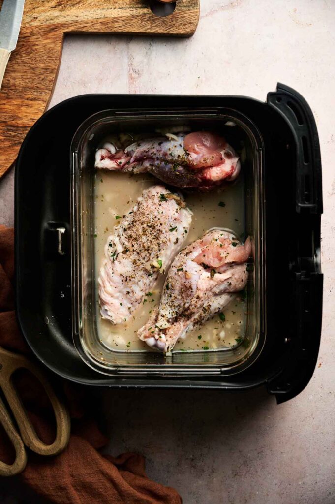 Air Fryer Turkey Wings seasoned with herbs and pepper are placed in the air fryer basket, ready for cooking. A cutting board and kitchen scissors are nearby for easy preparation.