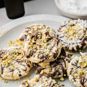 A plate of Air Fryer Cannoli Cookies topped with chocolate drizzle and crushed pistachios, with a bowl of powdered sugar in the background.
