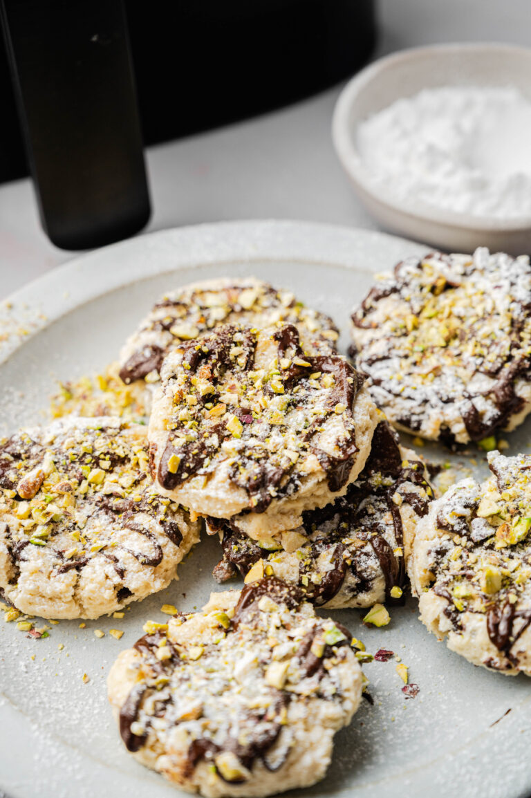 A plate of Air Fryer Cannoli Cookies topped with chocolate drizzle and crushed pistachios, with a bowl of powdered sugar in the background.