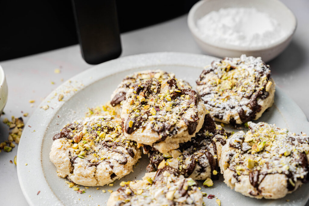 A plate of Air Fryer Cannoli Cookies topped with powdered sugar and chopped pistachios, with a bowl of powdered sugar in the background.