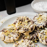A plate of Air Fryer Cannoli Cookies topped with powdered sugar and chopped pistachios, with a bowl of powdered sugar in the background.