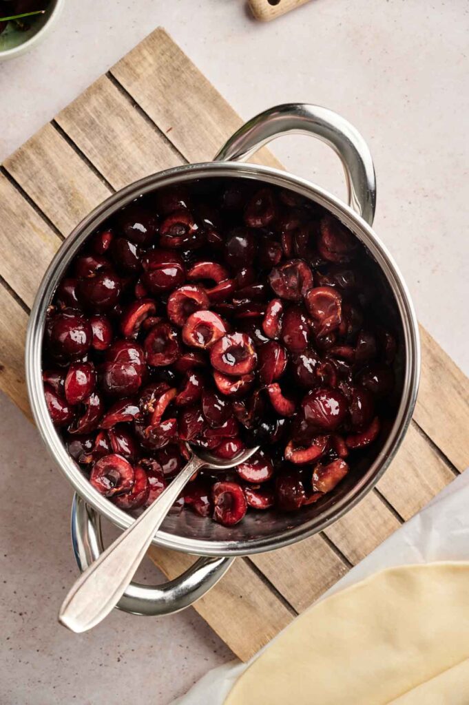 A stainless steel pot filled with sliced cherries sits on a wooden board, ready to become the filling for a homemade Air Fryer Cherry Pie, with a silver spoon resting inside.