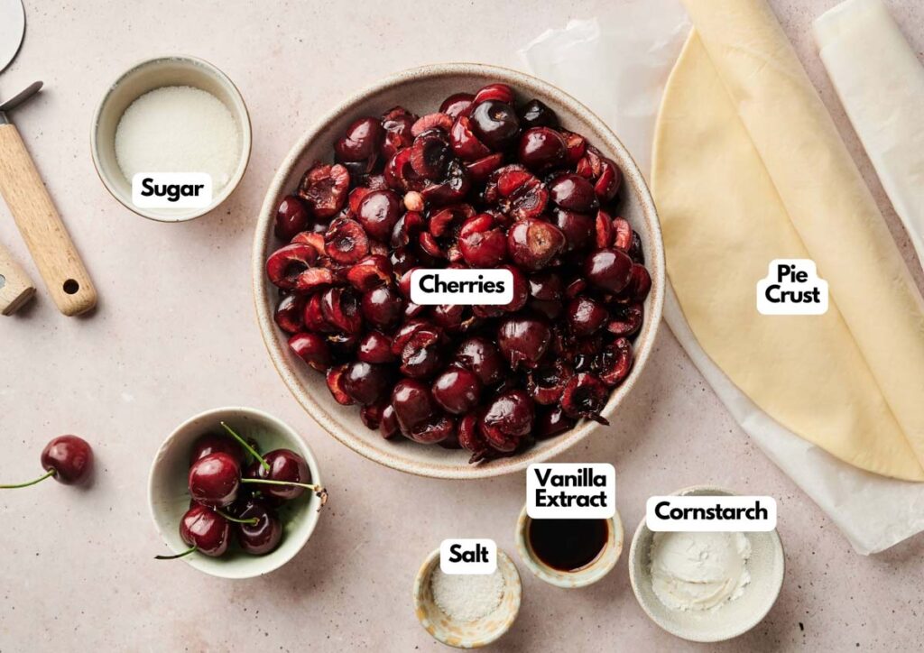 A bowl of cherries surrounded by labeled ingredients&mdash;sugar, pie crust, vanilla extract, salt, cornstarch, and fresh whole cherries&mdash;on a light countertop, perfect for making an Air Fryer Cherry Pie.