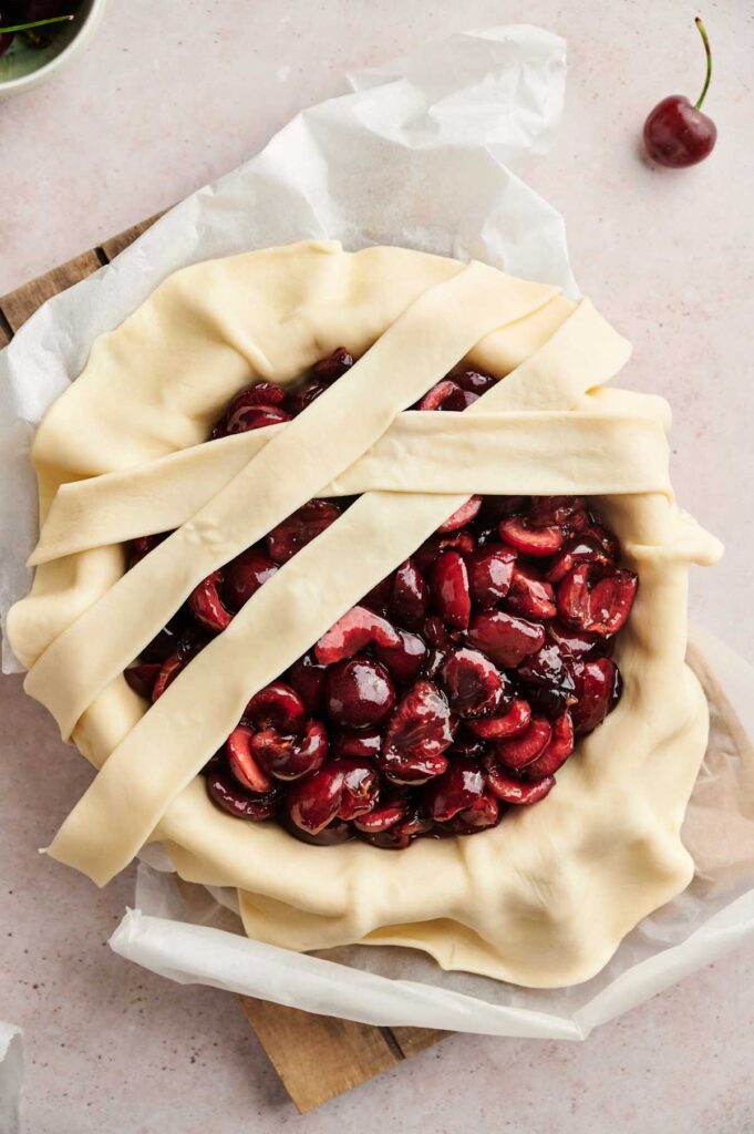 Unbaked Air Fryer Cherry Pie with lattice strips of dough partially placed over a filling of halved cherries in a parchment-lined pie dish on a light surface.