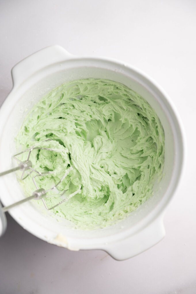 A mixing bowl containing light green whipped frosting with a handheld electric mixer resting inside&mdash;perfect for topping your Air Fryer Mint Chocolate Brownies.