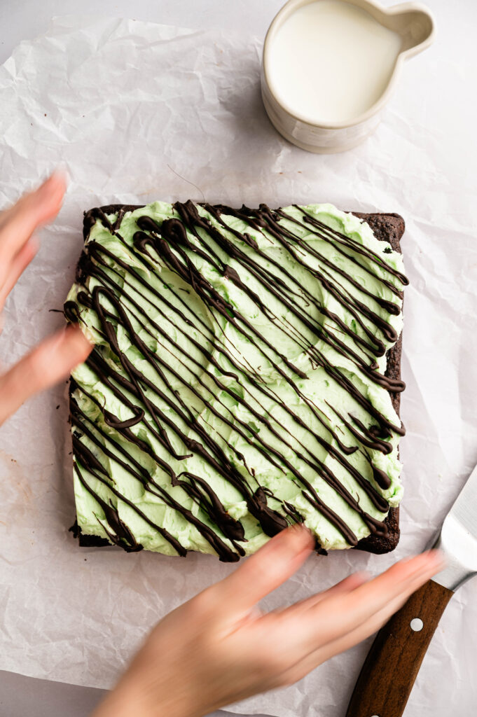 A square Air Fryer Mint Chocolate Brownie with green frosting and chocolate drizzle sits on parchment paper beside a knife and a small pitcher of milk. Hands are visible near the dessert.