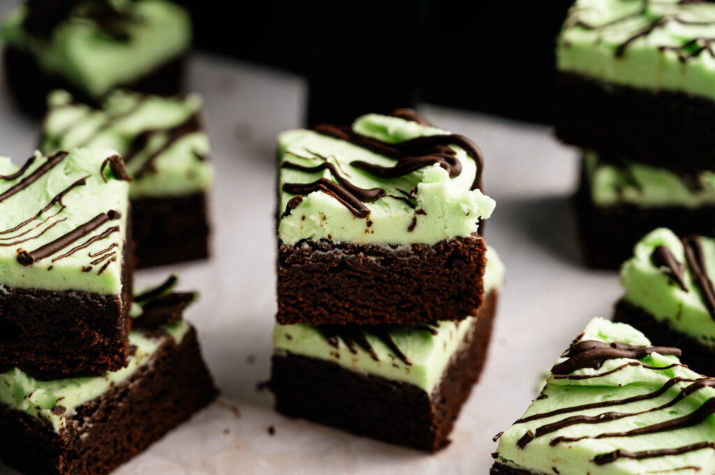 Air Fryer Mint Chocolate Brownies are stacked high, topped with mint green frosting and drizzled with chocolate, displayed beautifully on a white surface.