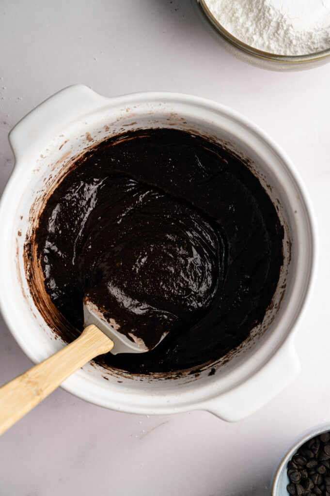 A white mixing bowl containing thick chocolate batter for Air Fryer Mint Chocolate Brownies with a spatula, shown on a light surface with bowls of flour and chocolate chips nearby.