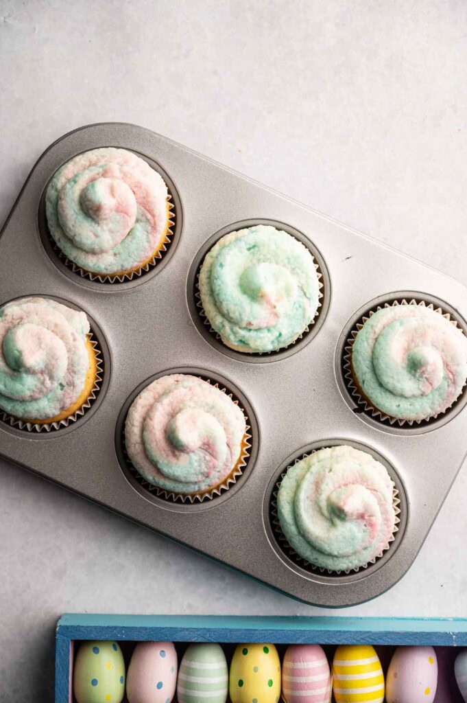 A muffin tin with six Easter Cupcakes, each pastel-frosted, sits above a box of colorful, decorated Easter eggs on a light surface.