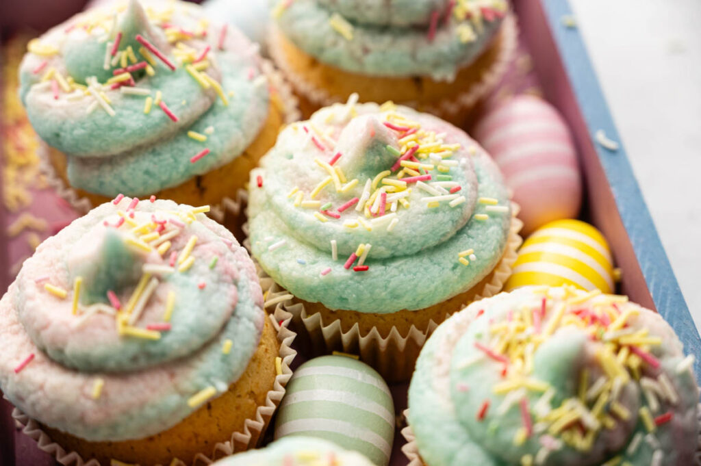 Easter Cupcakes with pastel blue and pink frosting topped with colorful sprinkles, surrounded by decorative striped and solid Easter eggs.