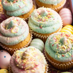 Easter Cupcakes with pastel-colored frosting and sprinkles are arranged in a box alongside decorated pastel Easter eggs.