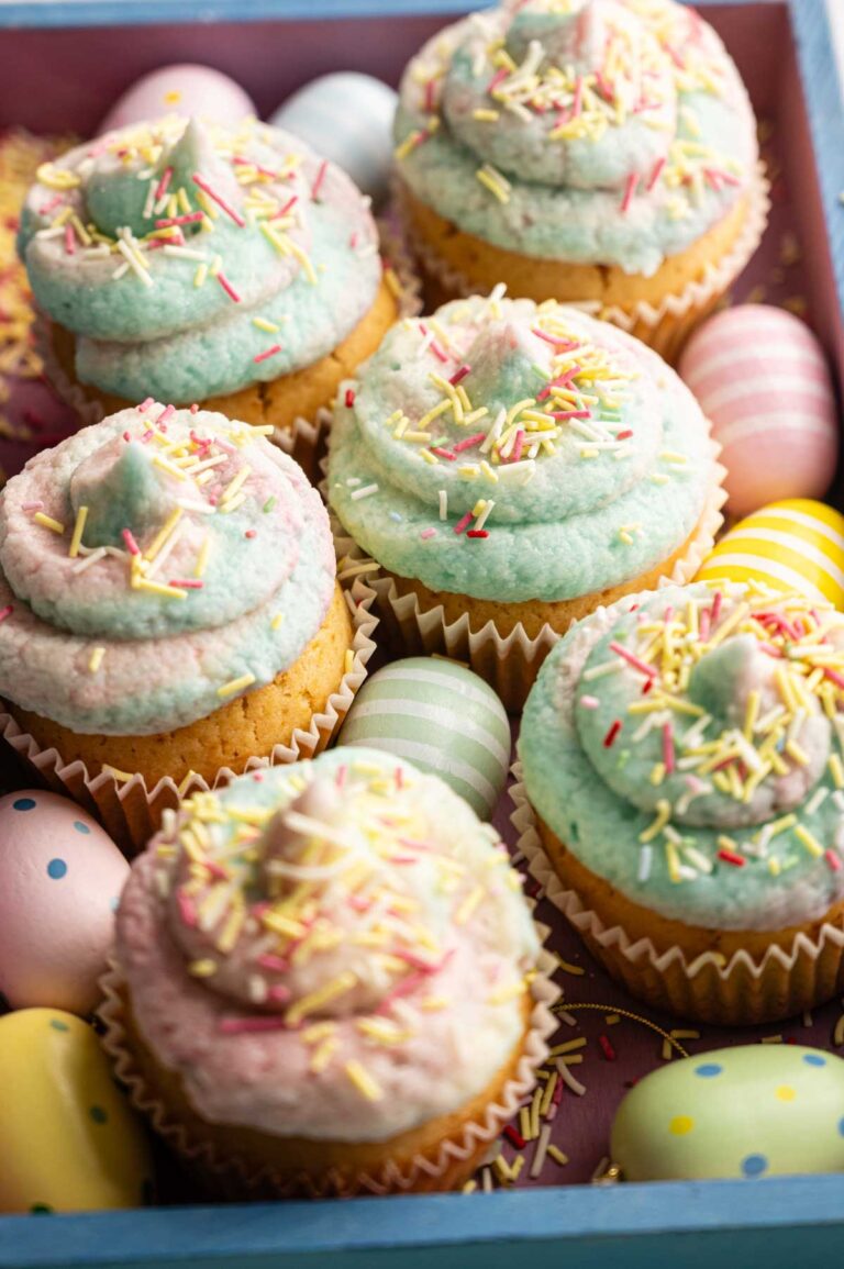 Easter Cupcakes with pastel-colored frosting and sprinkles are arranged in a box alongside decorated pastel Easter eggs.