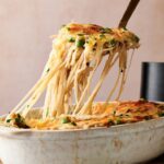 A serving spoon lifts cheesy baked spaghetti mixed with peas and herbs from a white casserole dish, reminiscent of comforting Air Fryer Chicken Tetrazzini.