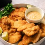 A plate of Air Fryer Crispy Chicken Thighs with lime wedges, a bunch of parsley, and a bowl of dipping sauce.
