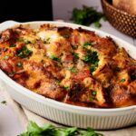An Air Fryer Eggs Benedict Casserole in a white dish, topped with melted cheese and chopped herbs, sits on a cloth with fresh parsley nearby.