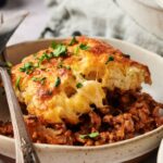 A plate of cheesy baked Air Fryer Sloppy Joe Casserole with a ground meat filling, topped with chopped parsley, served with a fork and knife.