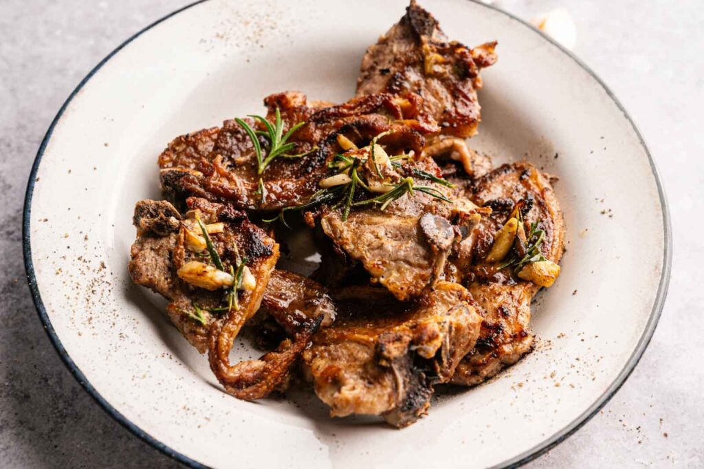 Plate of lamb loin chops garnished with fresh rosemary and roasted garlic, served on a white dish.