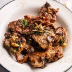 A white plate with several grilled lamb loin chops garnished with rosemary and sliced garlic.