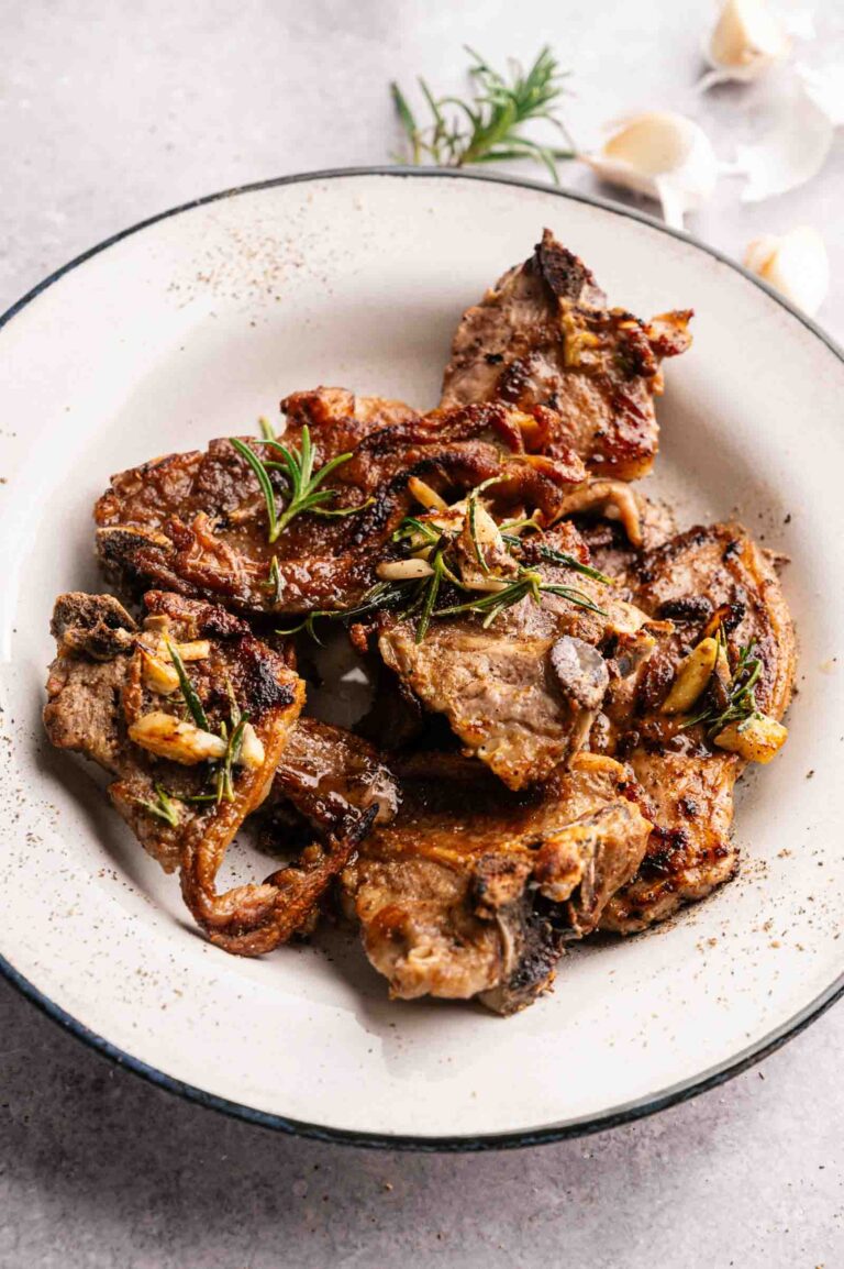 A white plate with several grilled lamb loin chops garnished with rosemary and sliced garlic.