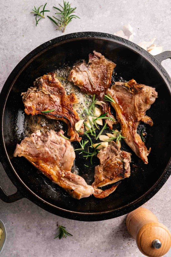 Lamb Loin Chops are being seared in a black skillet with fresh rosemary sprigs, sliced garlic, and melted butter.