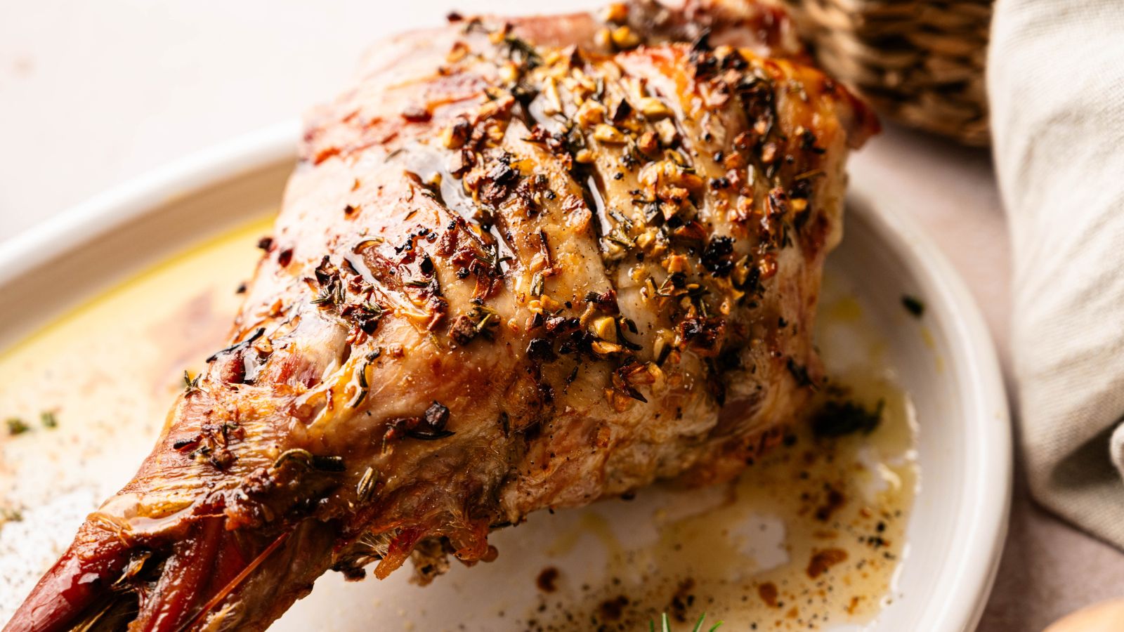 A cooked, herb-crusted leg of lamb rests on a white plate, garnished with seasonings and juices visible on the dish.