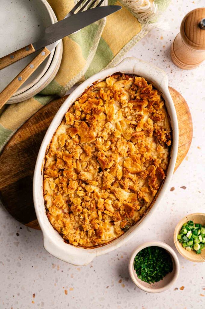 A Neiman Marcus Chicken Casserole with a golden, crumbly topping is shown in an oval dish on a wooden board, alongside chopped herbs and plates with utensils.