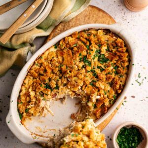 A Neiman Marcus Chicken Casserole with a golden, crumbly topping sits on a table; a serving has been scooped out, revealing the creamy interior. Plates, utensils, and seasonings are nearby.