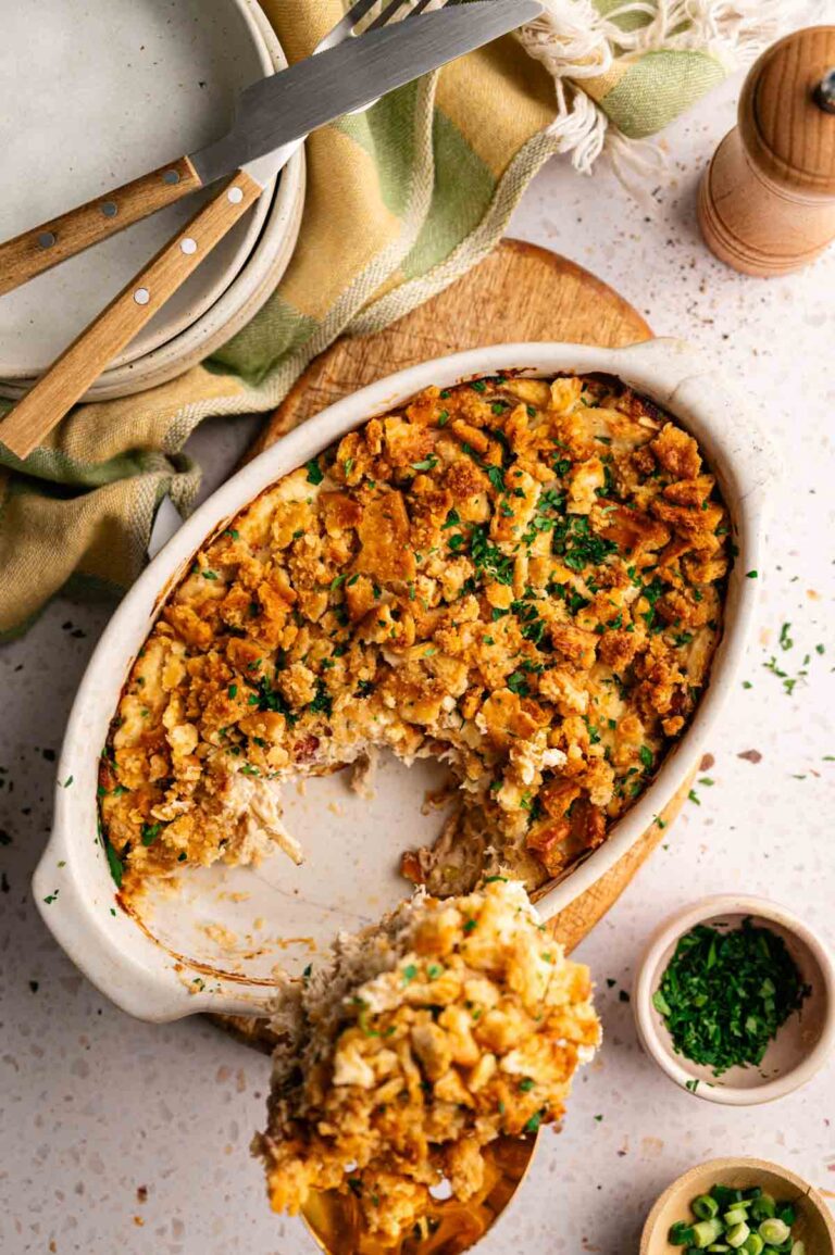 A Neiman Marcus Chicken Casserole with a golden, crumbly topping sits on a table; a serving has been scooped out, revealing the creamy interior. Plates, utensils, and seasonings are nearby.