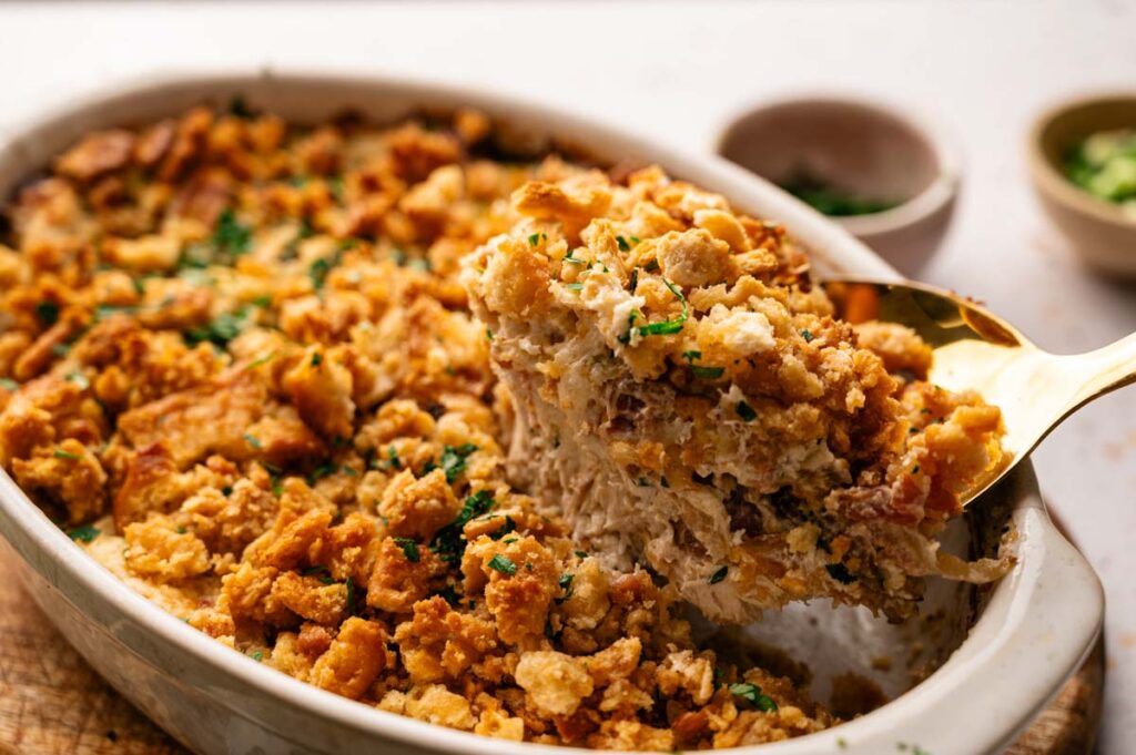 A casserole dish filled with a golden, crumb-topped Neiman Marcus Chicken Casserole, with a serving being lifted out on a spatula; chopped herbs are sprinkled on top.