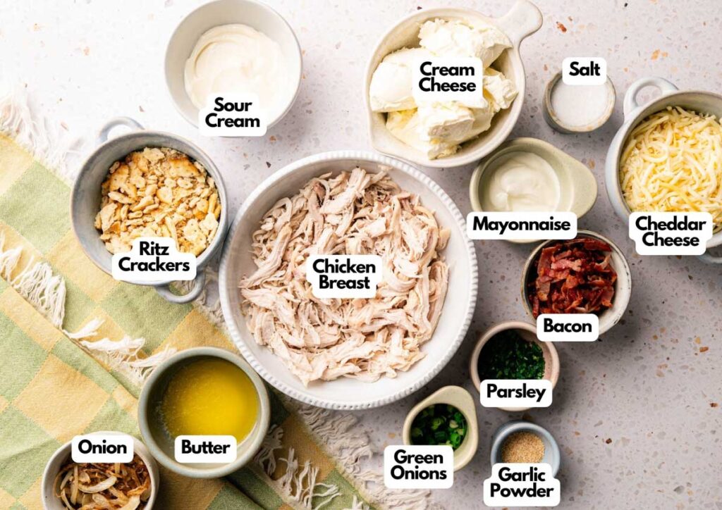 Top view of labeled ingredients in bowls for Neiman Marcus Chicken Casserole, including shredded chicken, sour cream, cream cheese, cheddar, mayonnaise, bacon, onion, Ritz crackers, butter, and seasonings.