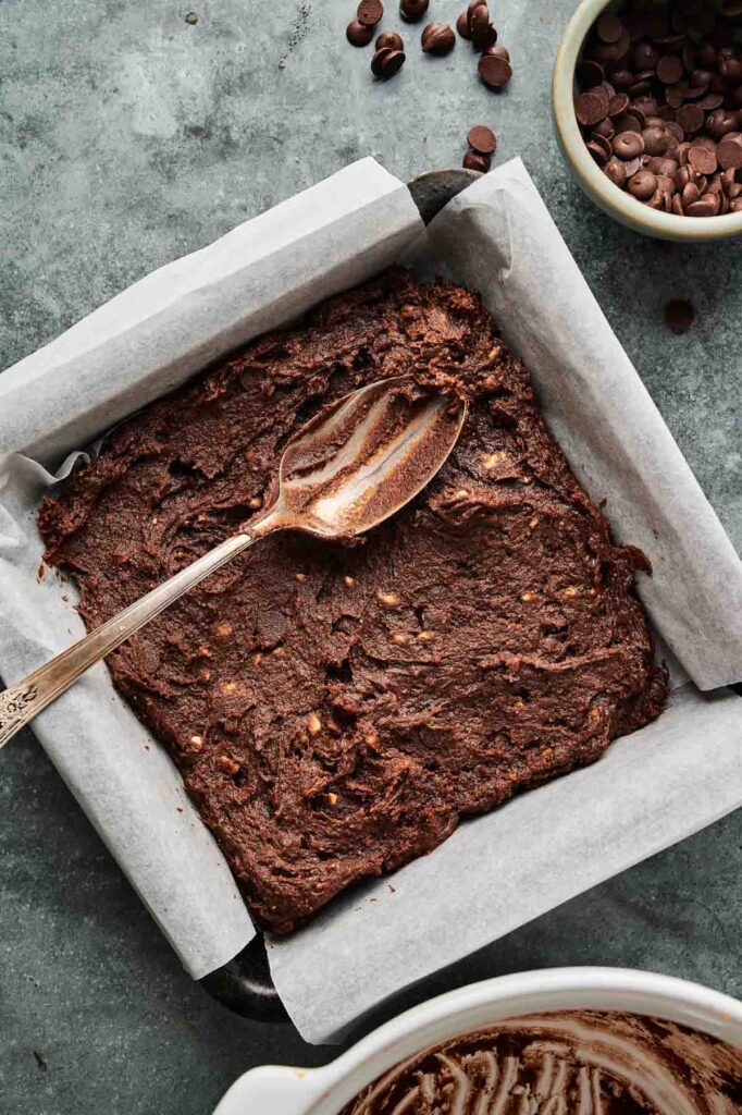 A square baking pan lined with parchment paper, filled with unbaked Air Fryer Sweet Potato Brownies batter and a spoon resting on top; chocolate chips and coffee beans are nearby.