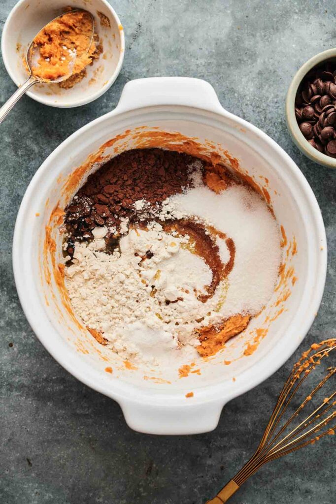 A white mixing bowl with flour, sugar, cocoa powder, and sweet potato sits on a counter next to a whisk, a bowl of chocolate chips&mdash;perfect for making Air Fryer Sweet Potato Brownies&mdash;and a small bowl with sweet potato residue and a spoon.