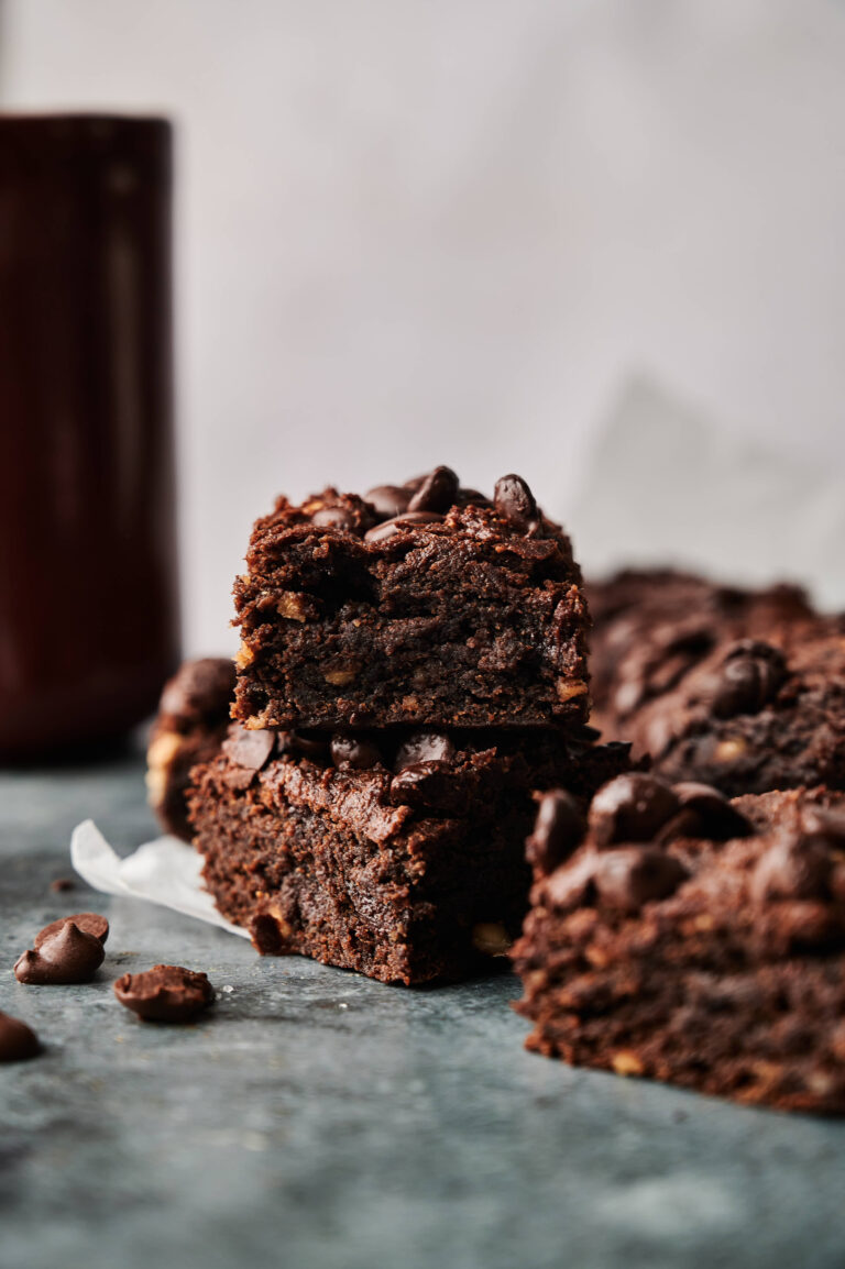 Stack of rich Air Fryer Sweet Potato Brownies with chocolate chips on a dark surface, surrounded by more brownies and scattered chocolate chips.
