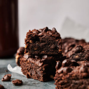 Stack of rich Air Fryer Sweet Potato Brownies with chocolate chips on a dark surface, surrounded by more brownies and scattered chocolate chips.