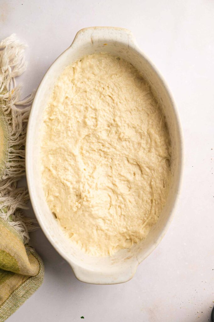 Oval ceramic dish filled with a smooth, pale batter or dough—perfect for preparing a classic John Wayne Casserole—with a textured towel partially visible on the left side.