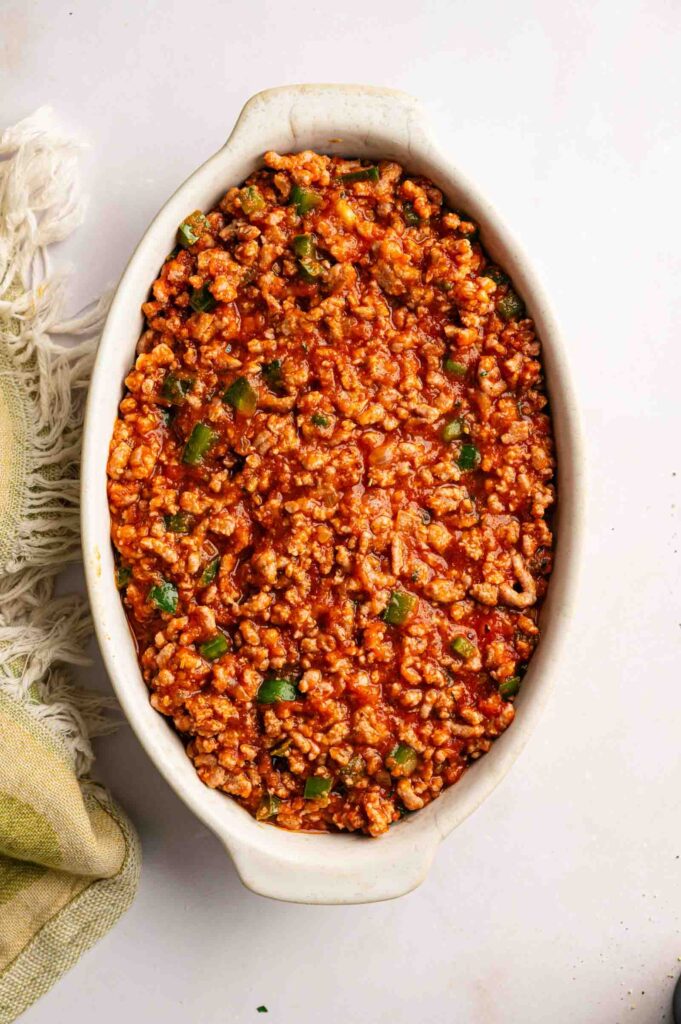Oval baking dish filled with cooked ground meat in tomato sauce and green bell pepper pieces, resembling a classic John Wayne Casserole, placed on a white surface next to a light-colored cloth.